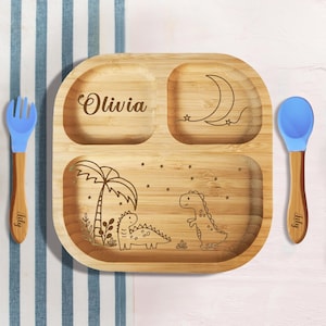 May include: A personalized wooden divided plate with a dinosaur and moon design. The plate is engraved with the name "Olivia". It comes with a blue silicone spoon and fork.