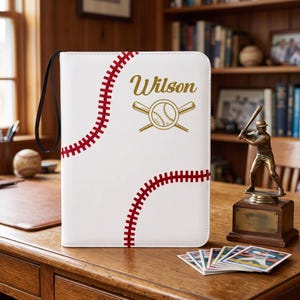 May include: A white rectangular binder featuring a red baseball stitch pattern across the front. The cover displays the name Wilson in gold script above a graphic of a baseball crossed with two bats. A black wrist strap is attached to the side.