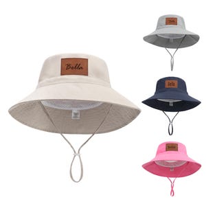 May include: A collection of sun hats in various colors: beige, gray, navy blue, and pink. Each hat features a wide brim, a chin strap, and a brown leather patch with the name "Bella" on the front. The hats are designed for sun protection.