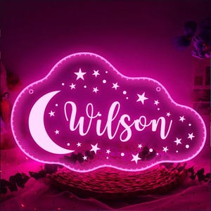 May include: A pink cloud-shaped LED night light with the name "Wilson" in a cursive font. The design includes a crescent moon and stars. The light is surrounded by a pink glow, creating a soft, ambient effect.