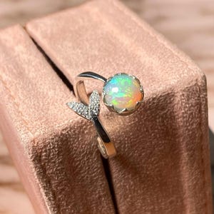 May include: A silver ring featuring a round opal gemstone and a small, diamond-encrusted mermaid tail design. The ring is displayed on a pink velvet box.
