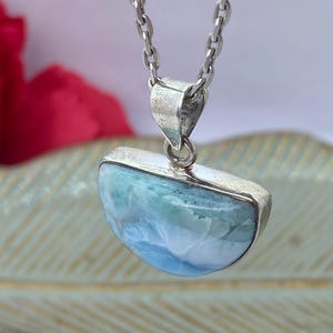 May include: A silver necklace featuring a pendant with a half-moon shaped, light blue and white gemstone. The gemstone is encased in a silver setting and hangs from a delicate silver chain. The background is blurred, with hints of red and gold.