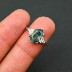 Puede incluir: Un anillo de plata con una piedra preciosa de ágata verde y blanca en un engaste hexagonal. El anillo está adornado con pequeñas piedras blancas.