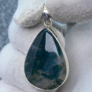 Puede incluir: Un colgante en forma de lágrima con un bisel plateado. El colgante presenta una piedra de ágata musgosa con patrones verdes, blancos y azules. La piedra está pulida y tiene un acabado brillante. El colgante está suspendido de una fianza plateada.