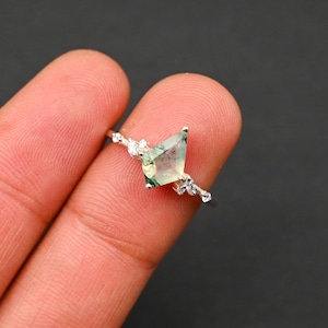 May include: A silver ring featuring a green moss agate gemstone and small clear stones. The ring is worn on a finger.
