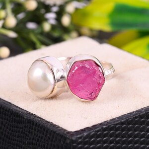 Pode incluir: Um anel de prata com uma gema rosa e uma pérola branca. O anel está engastado em uma banda de prata.