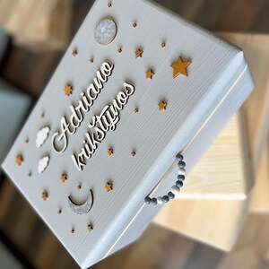 Puede incluir: Una caja de madera gris claro con un asa de cuentas. La caja está decorada con estrellas, nubes y lunas. El texto "Adriano krukstyno" está escrito en la caja.