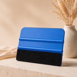 May include: A blue squeegee with a black felt edge rests on a stone block. The tool is designed for applying vinyl or window film. A vase with dried flowers is in the background.