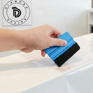Puede incluir: Una espátula azul y negra se utiliza para aplicar una película adhesiva transparente a una superficie blanca. El logotipo de Diamond Design es visible en la esquina superior izquierda. La película se está aplicando a una encimera.