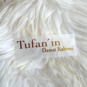 May include: A rectangular white sticker featuring the gold-colored text Tufan'ın Damat Kahvesi in a serif font. The sticker is placed against a background of long, white, soft-textured faux fur material with a shaggy appearance.