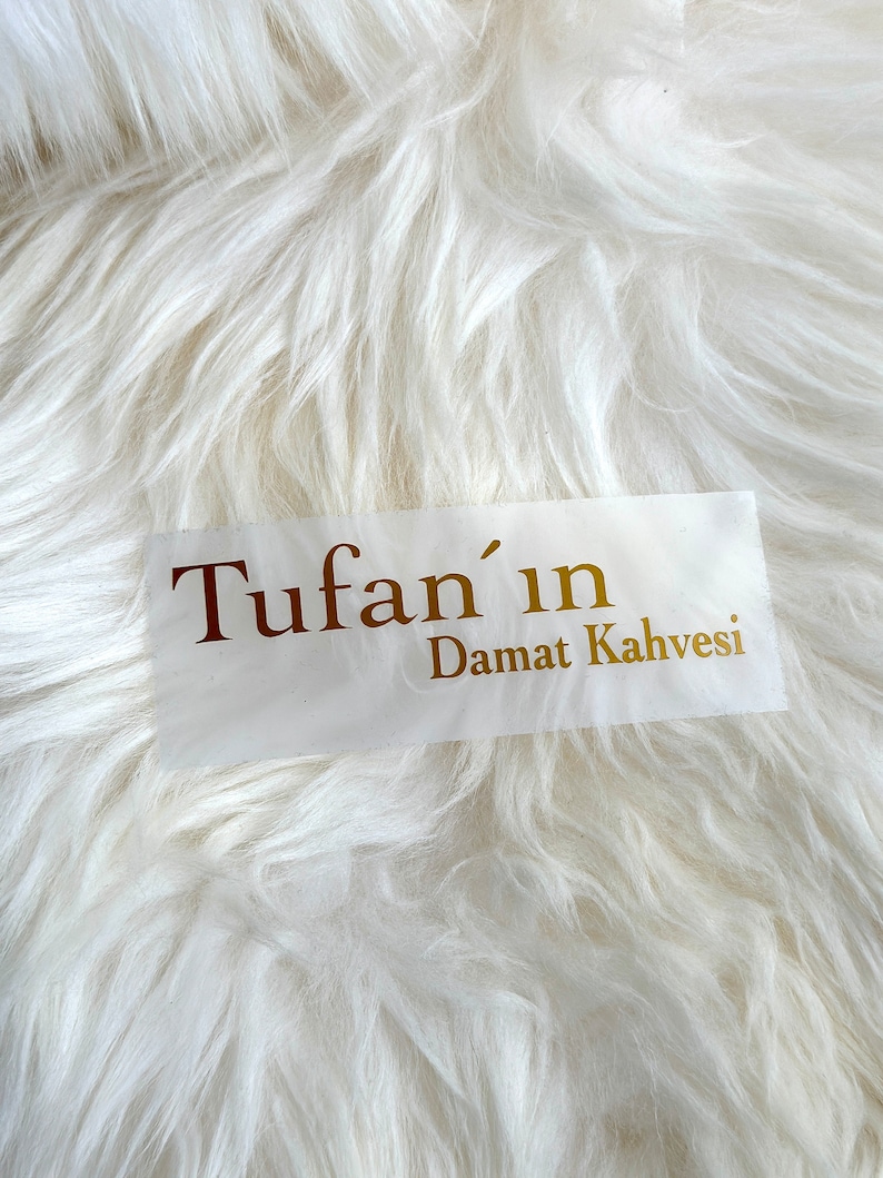 May include: A rectangular white sticker featuring the gold-colored text Tufan'ın Damat Kahvesi in a serif font. The sticker is placed against a background of long, white, soft-textured faux fur material with a shaggy appearance.