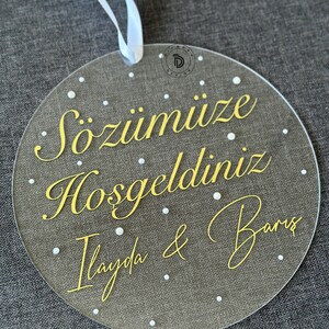 May include: A clear, round acrylic sign with gold lettering and white dots. The sign has the text "S&ouml;z&uuml;m&uuml;ze Hoşgeldiniz Ilayda & Barış". A white ribbon is attached at the top for hanging.