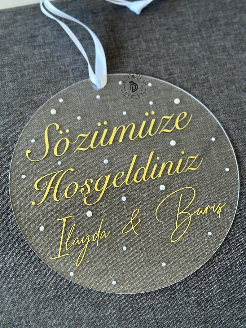 May include: A clear, round acrylic sign with gold lettering and white dots. The sign has the text "S&ouml;z&uuml;m&uuml;ze Hoşgeldiniz Ilayda & Barış". A white ribbon is attached at the top for hanging.