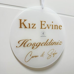 Pu&ograve; includere: Un cartello rotondo bianco con lettere dorate che recita "Kız Evine Hoşgeldiniz Ceren & Sefer".