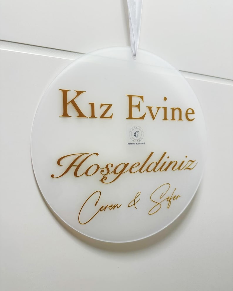 Pu&ograve; includere: Un cartello rotondo bianco con lettere dorate che recita "Kız Evine Hoşgeldiniz Ceren & Sefer".