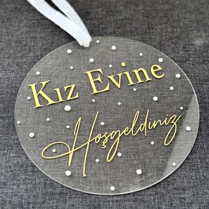 Pu&ograve; includere: Un ornamento rotondo in acrilico trasparente con lettere dorate che recitano "Kız Evine Hoşgeldiniz", che si traduce in "Benvenuti nella casa della ragazza". L'ornamento ha punti bianchi attorno ai bordi.