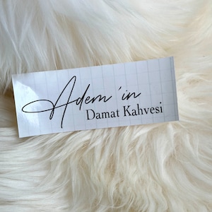 May include: A rectangular white sticker with a faint light gray grid pattern. The text "Adem'in" is printed in a black cursive script font, followed by the text "Damat Kahvesi" in a simple, clean black sans-serif typeface.