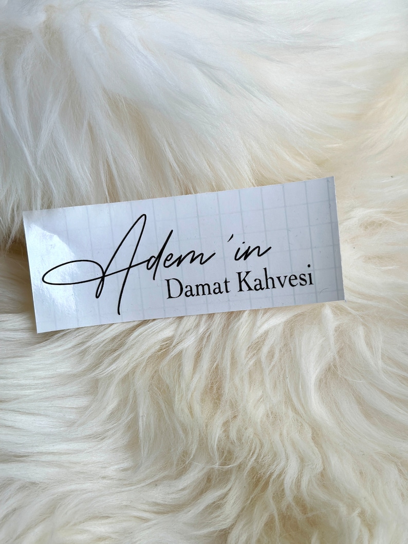 May include: A rectangular white sticker with a faint light gray grid pattern. The text "Adem'in" is printed in a black cursive script font, followed by the text "Damat Kahvesi" in a simple, clean black sans-serif typeface.