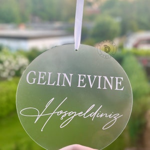 May include: A round, clear acrylic sign with the text "GELIN EVINE Hosgeldiniz" in white lettering. The sign is hanging from a white ribbon.