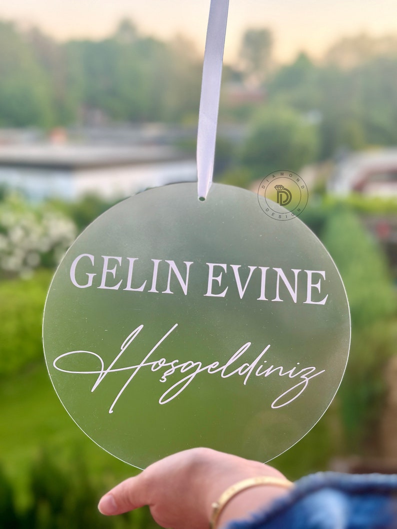 May include: A round, clear acrylic sign with the text "GELIN EVINE Hosgeldiniz" in white lettering. The sign is hanging from a white ribbon.