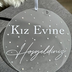 May include: A clear, round decorative sign with the text "Kız Evine Hoşgeldiniz" in white. The sign is adorned with small, scattered, iridescent dots and has a light blue ribbon for hanging. A logo is visible in the upper right corner.