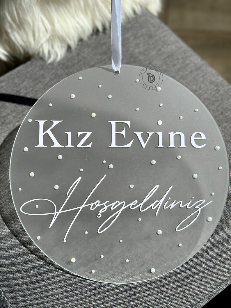 May include: A clear, round decorative sign with the text "Kız Evine Hoşgeldiniz" in white. The sign is adorned with small, scattered, iridescent dots and has a light blue ribbon for hanging. A logo is visible in the upper right corner.