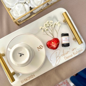 May include: A rectangular white serving tray with gold handles, featuring the text "Bekir'in Damat Kahvesi" and "01|06|2024". It holds a white coffee cup and saucer with "AB" monogram, a red heart-shaped chocolate, and a small sprig of white baby's breath.