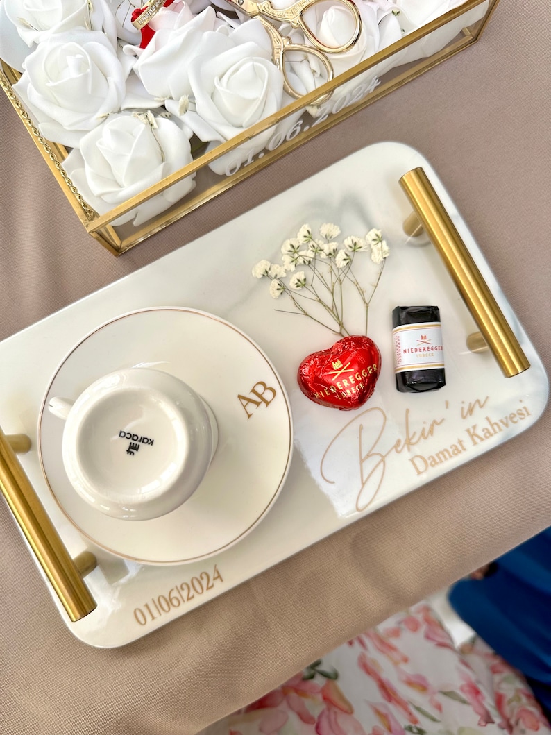 May include: A rectangular white serving tray with gold handles, featuring the text "Bekir'in Damat Kahvesi" and "01|06|2024". It holds a white coffee cup and saucer with "AB" monogram, a red heart-shaped chocolate, and a small sprig of white baby's breath.
