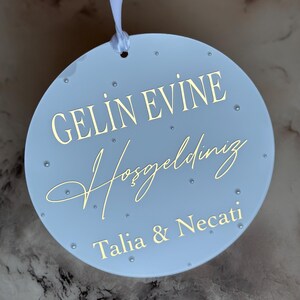 May include: A light blue, round decorative sign with gold lettering and small, clear embellishments. The sign reads "GELIN EVINE Hosgeldiniz Talia & Necati" and is suspended by a white ribbon. A wedding or housewarming gift.