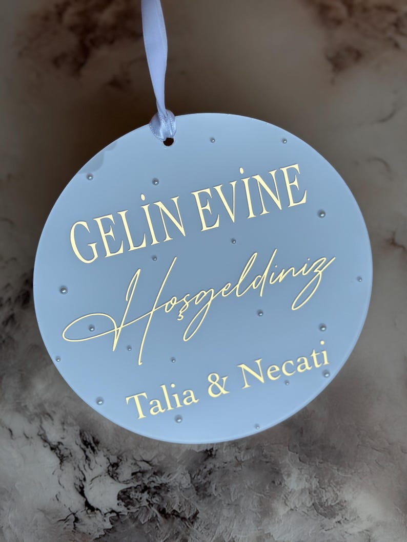 May include: A light blue, round decorative sign with gold lettering and small, clear embellishments. The sign reads "GELIN EVINE Hosgeldiniz Talia & Necati" and is suspended by a white ribbon. A wedding or housewarming gift.