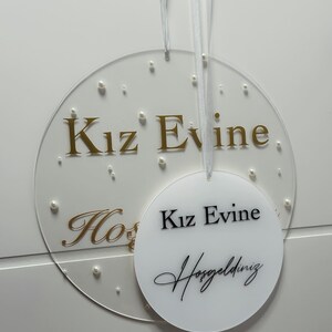 Pu&ograve; includere: Due cerchi in acrilico bianco con lettere dorate. Il cerchio pi&ugrave; grande ha il testo "Kiz Evine" in lettere dorate e piccoli punti bianchi. Il cerchio pi&ugrave; piccolo ha il testo "Kiz Evine" in lettere nere e il testo "Hosgeldiniz" in lettere corsive nere.