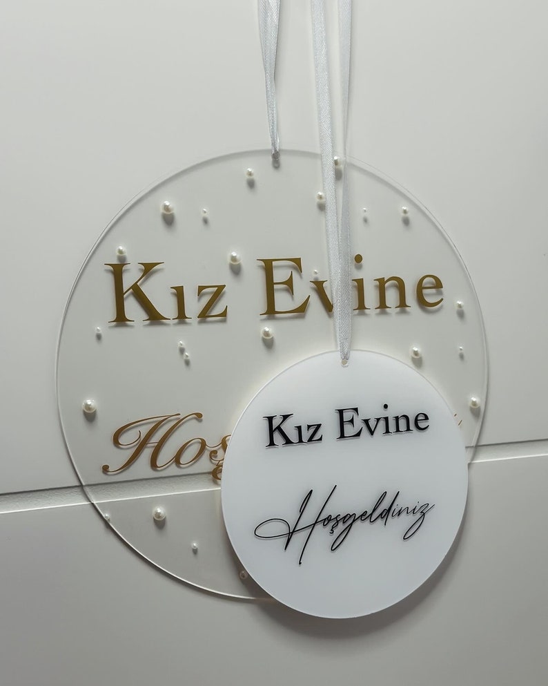 Pu&ograve; includere: Due cerchi in acrilico bianco con lettere dorate. Il cerchio pi&ugrave; grande ha il testo "Kiz Evine" in lettere dorate e piccoli punti bianchi. Il cerchio pi&ugrave; piccolo ha il testo "Kiz Evine" in lettere nere e il testo "Hosgeldiniz" in lettere corsive nere.