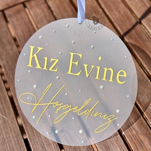 May include: A round, gray sign with the text "Kız Evine Hoşgeldiniz" in yellow script. The sign is decorated with small, white pearl-like embellishments and has a white ribbon for hanging.
