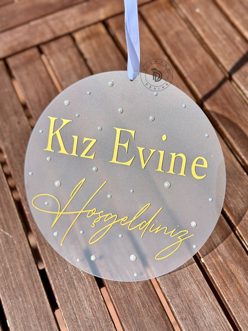 May include: A round, gray sign with the text "Kız Evine Hoşgeldiniz" in yellow script. The sign is decorated with small, white pearl-like embellishments and has a white ribbon for hanging.