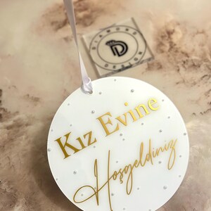 Pu&ograve; includere: Un cartello rotondo bianco con lettere dorate che dice "Kiz Evine Hosgeldiniz". Il cartello &egrave; decorato con piccoli punti bianchi.