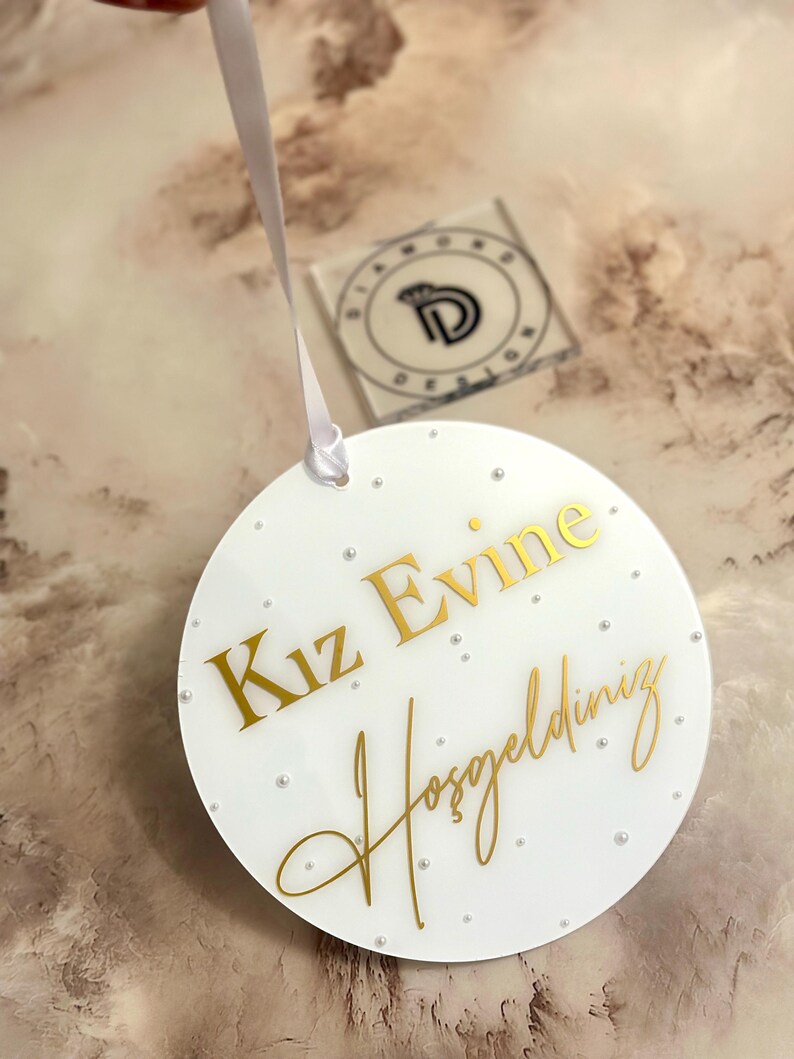 Pu&ograve; includere: Un cartello rotondo bianco con lettere dorate che dice "Kiz Evine Hosgeldiniz". Il cartello &egrave; decorato con piccoli punti bianchi.