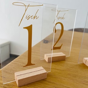 Könnte beinhalten: Drei transparente Acryl-Tischnummern-Halter mit goldener Beschriftung. Die Nummern sind 1 und 2. Die Halter stehen auf einem Holzsockel.