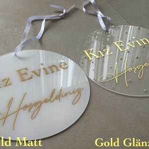 May include: Two oval-shaped signs with white ribbons for hanging. One is opaque white with gold text, the other is clear with gold text and small decorative dots. The text reads "Kız Evine Hoşgeldiniz".