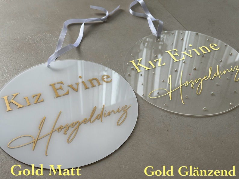 May include: Two oval-shaped signs with white ribbons for hanging. One is opaque white with gold text, the other is clear with gold text and small decorative dots. The text reads "Kız Evine Hoşgeldiniz".