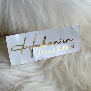 May include: A rectangular white adhesive label featuring a faint grid pattern and gold metallic lettering that reads Hakan'ın Damat Kahvesi. The text is presented in a combination of a cursive script font and a clean sans-serif typeface.
