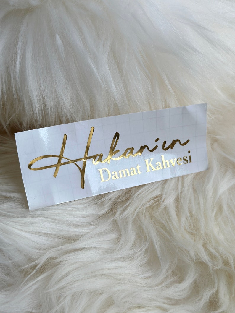 May include: A rectangular white adhesive label featuring a faint grid pattern and gold metallic lettering that reads Hakan'ın Damat Kahvesi. The text is presented in a combination of a cursive script font and a clean sans-serif typeface.