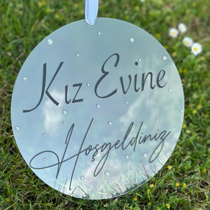 May include: A round, clear acrylic sign with the text "Kız Evine Hoşgeldiniz" in black lettering. The sign is decorated with small, white rhinestones.