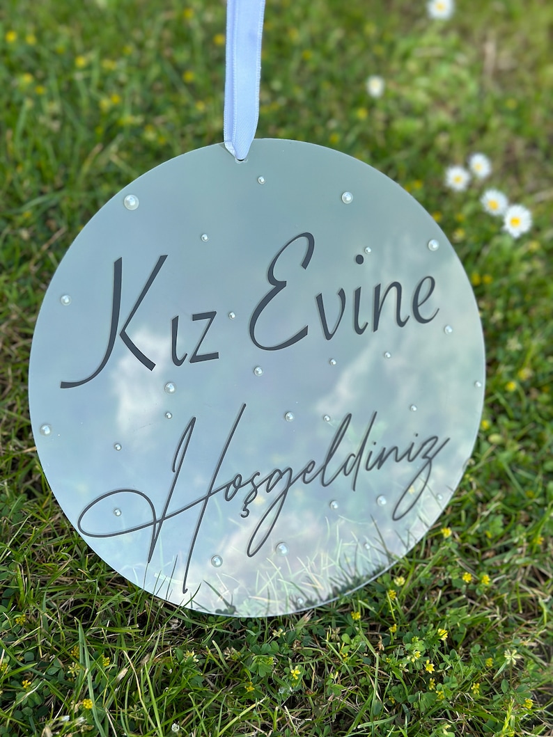 May include: A round, clear acrylic sign with the text "Kız Evine Hoşgeldiniz" in black lettering. The sign is decorated with small, white rhinestones.