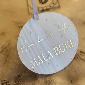 May include: A white round ornament with a light blue background and gold lettering. The ornament has a white ribbon attached to the top. The text on the ornament reads "Hun bi xer hatin MALA BUKE".