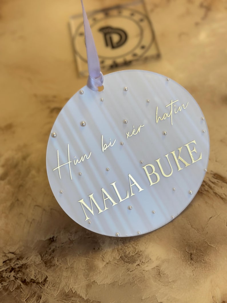 May include: A white round ornament with a light blue background and gold lettering. The ornament has a white ribbon attached to the top. The text on the ornament reads "Hun bi xer hatin MALA BUKE".