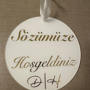 May include: White circular ornament with gold lettering and a white ribbon. The ornament has the words "S&ouml;z&uuml;m&uuml;ze Hoşgeldiniz" and the initials "D|H" in a decorative font. The background is a neutral tan fabric.