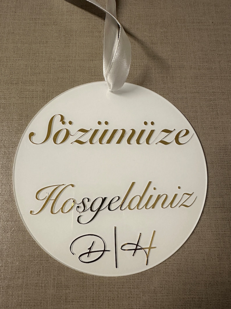 May include: White circular ornament with gold lettering and a white ribbon. The ornament has the words "S&ouml;z&uuml;m&uuml;ze Hoşgeldiniz" and the initials "D|H" in a decorative font. The background is a neutral tan fabric.