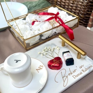 May include: A rectangular white marble-look serving tray with gold handles, featuring the text 'Bekir' in Damat Kahvesi' and '01/06/2024'. It holds a white coffee cup, a heart-shaped red chocolate, a small rectangular chocolate, and dried flowers.