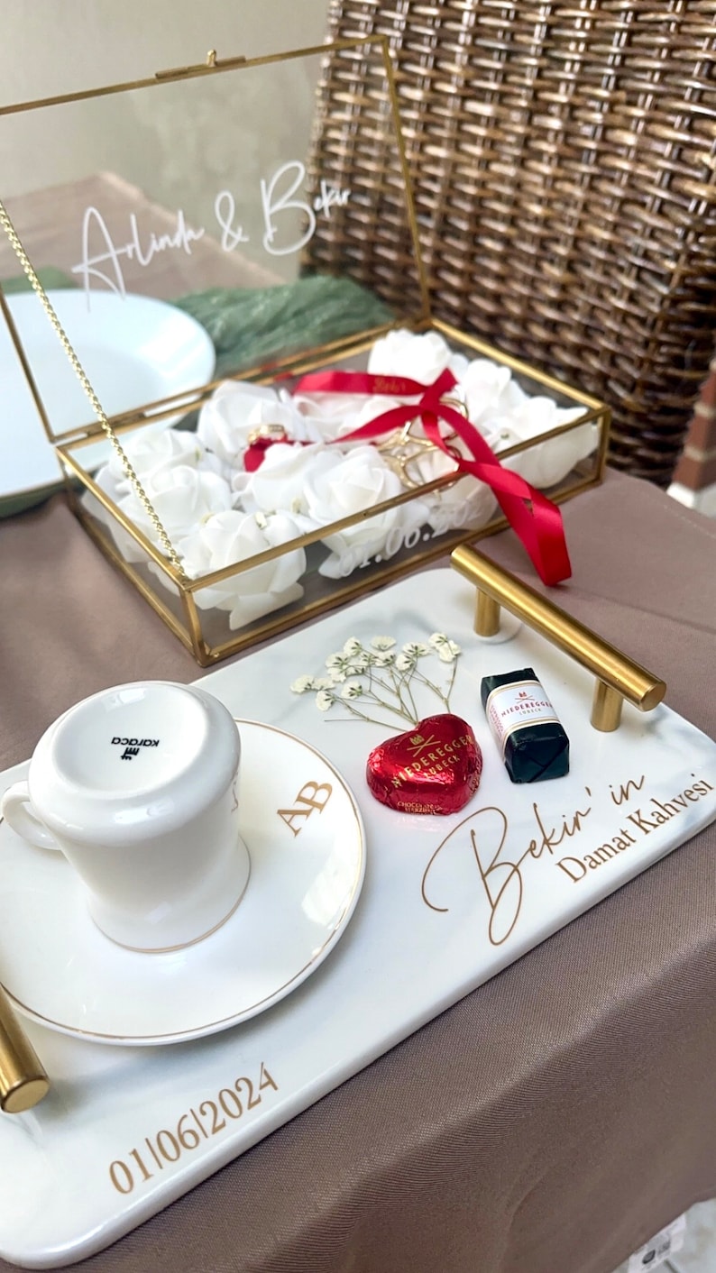 May include: A rectangular white marble-look serving tray with gold handles, featuring the text 'Bekir' in Damat Kahvesi' and '01/06/2024'. It holds a white coffee cup, a heart-shaped red chocolate, a small rectangular chocolate, and dried flowers.