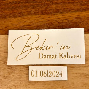 May include: A rectangular white label with gold text reading Bekir'in Damat Kahvesi in a mix of script and serif fonts. Below it, a smaller white label features the gold date 01|06|2024 in a clean serif font. Both labels are placed on a wood surface.